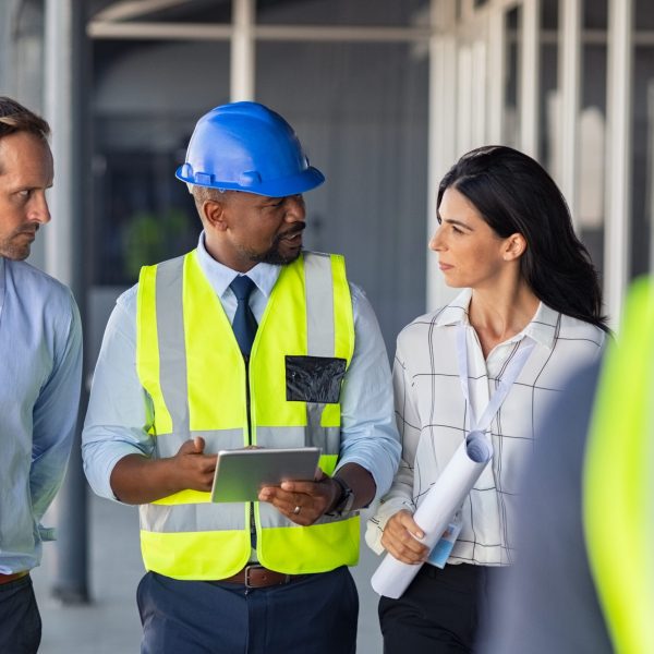 Team of construction workers discussing project details with executive supervisor holding blueprints and digital tablet. Team of architects and civil engineers inspecting construction site. Structural african engineer and architect with work vests discuss the construction process.